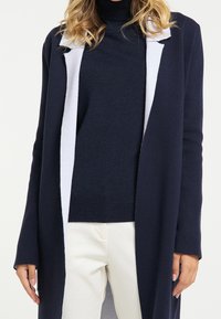 Marineblauer Cardigan mit weißem Akzent entlang der Vorderseite, weicher Textur, offenem Design, langen Ärmeln und locker sitzend über einem Rollkragenpullover.