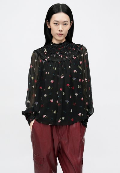Blusa nera trasparente con colletto alto arricciato, decorata con motivi floreali colorati. Maniche lunghe con polsini arricciati. Abbinata a pantaloni bordeaux.