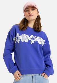 Sudadera azul con diseño floral blanco en el pecho. Cuello redondo, mangas largas y puños acanalados. Combinada con una gorra rosa claro.