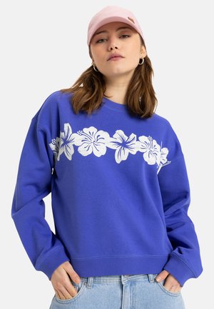 Sudadera azul con diseño floral blanco en el pecho. Cuello redondo, mangas largas y puños acanalados. Combinada con una gorra rosa claro.