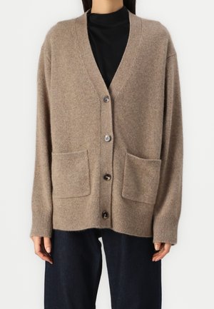 Mujer con un cárdigan beige suelto con botones al frente, dos bolsillos y una camiseta negra debajo, combinada con pantalones oscuros.