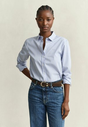 Femme aux cheveux tressés portant une chemise à rayures bleues et blanches boutonnée, rentrée dans un jean bleu avec une ceinture marron, debout devant un fond uni.