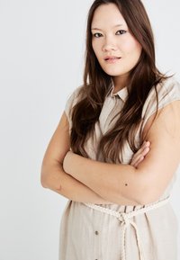 Vestido de linho bege com decote em colarinho, mangas curtas e fecho de botão à frente. Cinto trançado branco na cintura. O modelo tem cabelo longo e escuro.