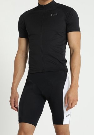 Homme portant un maillot de cyclisme à manches courtes noir et un cuissard de cyclisme noir avec des panneaux latéraux blancs, tous deux de la marque "Gore".