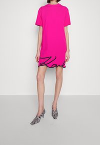 Helles pinkes Kurzarmkleid mit rundem Halsausschnitt, versehen mit einem schwarzen, geschwungenen "Karl"-Logo am Saum. Model trägt Schuhe mit Zebramuster.