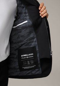 Czarna kurtka o gładkiej powierzchni, podszewka w wzorzysty szary wzór. Zawiera metkę z opisem cech, takich jak elastyczność i szybkie schnięcie.