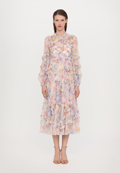 Robe transparente à motifs floraux, à manches longues, avec des accents volants, dans des couleurs pastel et une jupe à étages. Portée avec des talons nus à lanières.