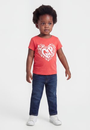 Ungt barn med krøllete hår iført en korallfarget t-skjorte med et blomsterhjerte og teksten "GAP", mørke jeans og hvite joggesko, stående på hvit bakgrunn.