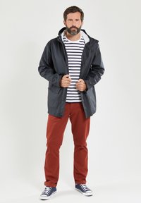 Zwart waterdicht herenjack met capuchon, voorzien van een ritssluiting aan de voorkant en twee zakken, gelegd over een wit en marineblauw gestreept shirt en rode broek.