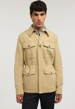 Giovane uomo con capelli scuri che indossa una giacca beige con bottoni e quattro tasche frontali sopra una camicia blu con colletto, in piedi davanti a uno sfondo semplice.