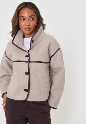 Mujer que lleva una chaqueta de forro polar beige con textura, con ribete oscuro y gran cuello, combinada con pantalones oscuros, posando sobre un fondo claro.