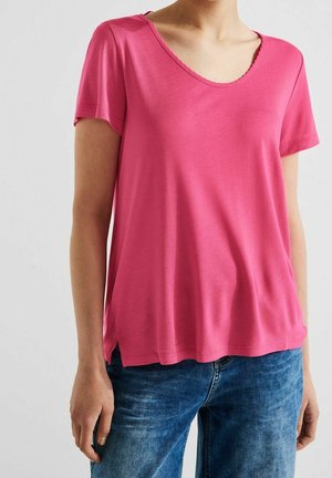 Vrouw draagt een loszittend, roze T-shirt met korte mouwen en een ronde hals, gecombineerd met een blauwe spijkerbroek, tegen een effen achtergrond.