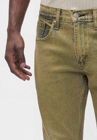 Gros plan de la main d’une personne reposant à côté d’un jean en denim vert olive délavé avec des coutures visibles et un bouton en métal.