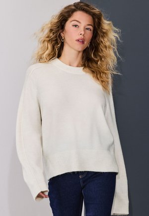 Giovane donna con capelli biondi ricci che indossa orecchini a cerchio dorati, un maglione bianco ampio e jeans blu scuro su uno sfondo a due tonalità di grigio.