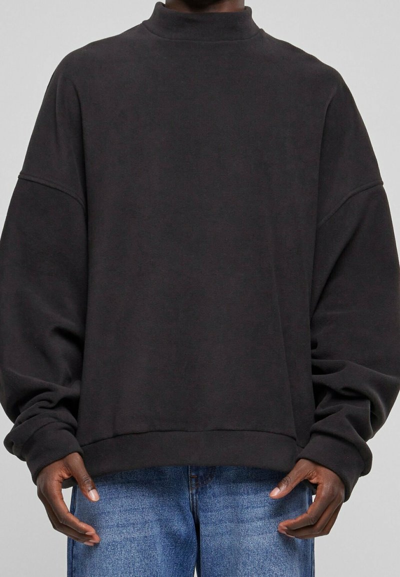 Persona con una sudadera negra holgada de manga larga y jeans de mezclilla azul, de pie frente a un fondo gris claro liso.