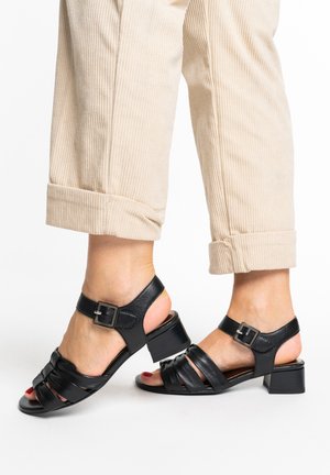 Voeten met beige corduroy broeken en zwarte leren sandaaltjes met hak, enkelriempjes en zilveren gespen op witte achtergrond.