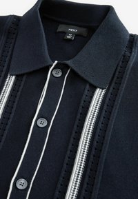 Navy blue knit polo shirt with black buttons, white vertical stripes, and textured patterns along the sides of the placket.