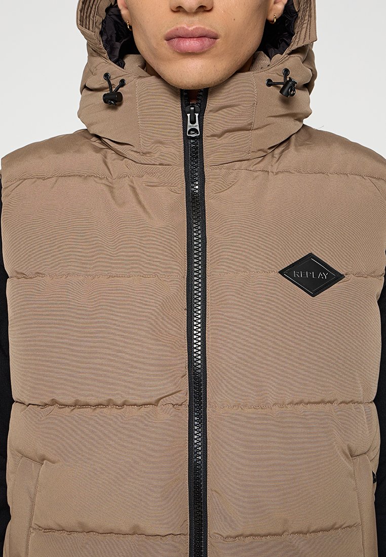 Braun gepolsterte Weste mit Reißverschluss vorne, hohem Kragen und verstellbarem Kapuzen. Enthält ein schwarzes Logo-Patch und Seitentaschen. Strukturiertes Material.