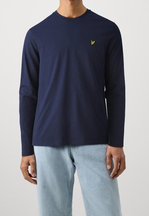 Mand iført marineblå langærmet skjorte med lille gul fuglelogo på brystet og lyseblå jeans, stående foran en ensfarvet baggrund.
