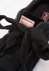 Gros plan d'une basket Puma noire avec des lacets noirs épais et texturés noués en nœud et un écusson rose avec le logo Puma cousu sur la languette.