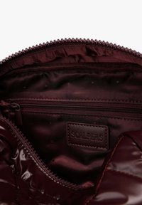 Interior burdeos de un bolso con un bolsillo con cremallera y una etiqueta de cuero con marca, con una textura exterior brillante y acolchada y un forro de tela suave.