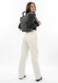 Sac à dos vert foncé en texture textile, avec deux poignées supérieures, des bretelles réglables et des poches zippées. Porté sur un haut noir et un pantalon crème.