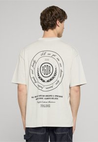 Světlé béžové tričko s černým kulat�ým obrázkem na zádech, který obsahuje text a motiv vavřínového věnce. Krátké rukávy a volný střih.