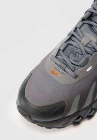 Pilkas sportinis batelis su juodu padu, tinklelio ir zomšinės viršutine dalimi, oranžiniu "swoosh" logotipu šalia raiščių ir banguoto pado dizainu.