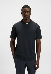 Man in een zwart T-shirt met korte mouwen met "BOSS" op de kraag, staand tegen een effen lichtgrijze achtergrond.