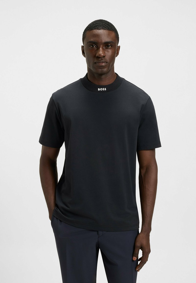 Man in een zwart T-shirt met korte mouwen met "BOSS" op de kraag, staand tegen een effen lichtgrijze achtergrond.
