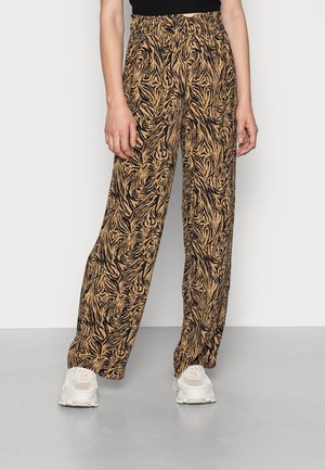 Kobieta w luźnych brązowych spodniach z czarnym, abstrakcyjnym zwierzęcym wzorem i białych trampkach, stojąca na tle jasnego, jednolitego tła.