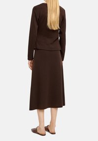 Kobieta z długimi blond włosami, ubrana w ciemno brązową bluzkę z długim rękawem i pasującą midi spódnicę, widziana z tyłu, w brązowych butach na płaskiej podeszwie.