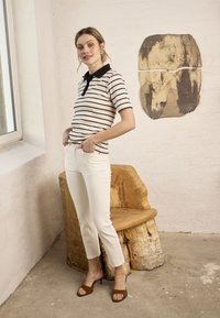 Femme portant un polo rayé et un pantalon blanc, debout pieds nus à côté d'une chaise en bois naturel face à un mur en briques blanches.
