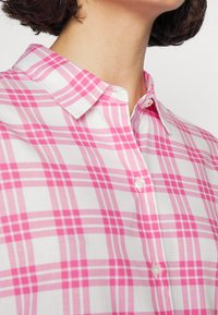 Růžová a bílá kostkovaná košile na knoflíky s špičatým límcem, vyrobená z lehkého materiálu a hladké textury. Knoflíky jsou bílé a kulaté.