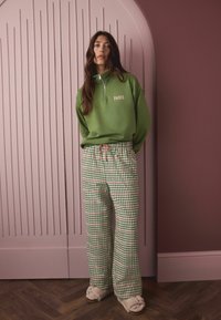 Green sweatshirt with “PARIS” text, zip neck; plaid pants in green, red, white; soft beige slippers; backdrop features pink paneled wall.
