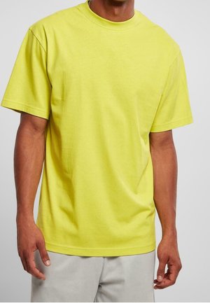 Homme portant un t-shirt jaune vif uni à manches courtes et un short gris clair sur un fond uni.