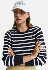 Gestreifter, marineblauer und weißer Rippstrickpullover mit rundem Halsausschnitt und engen Bündchen, kombiniert mit einer weißen Kappe mit blauer Schrift und Grafiken.