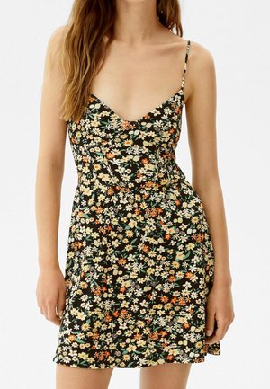 Mujer con un vestido negro sin mangas con estampado floral, tirantes finos y escote en V, de pie frente a un fondo blanco liso.