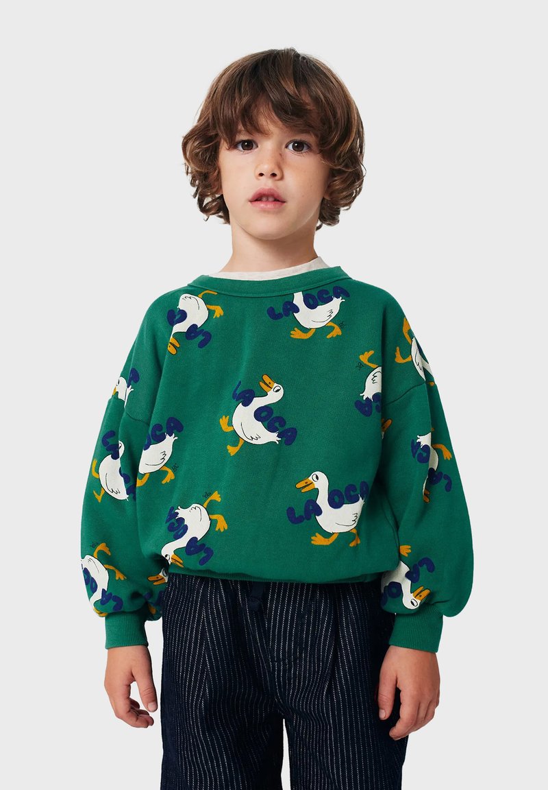 Felpa verde con motivo anatra e testo "LA OCA", con vestibilità rilassata e polsini a costine. Abbinata a pantaloni a righe blu navy.