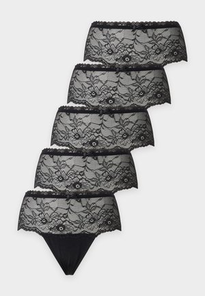 Mujer de pie descalza, con sujetador azul y ropa interior negra con encaje, sobre un fondo blanco liso.