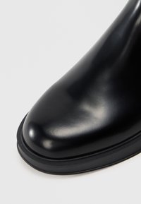 Bottine en cuir noir avec une surface lisse et brillante, un bout rond et des détails en caoutchouc noir contrastants sur la semelle.