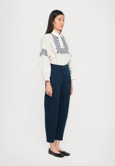 Blusa branca com padrões azuis, apresentando mangas bufantes e bordado no decote. Calças de cintura alta azul-escuro, com textura matte, e sapatos planos.