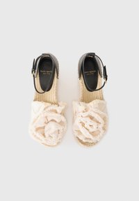 Espadrilles beige con un detalle floral texturizado en la parte frontal, tira negra en el tobillo y suela de yute trenzado. Marca dorada visible en el interior.