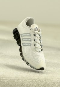 White Adidas running shoe with black segmented sole, mesh upper, three side stripes, and laces on a soft beige surface.