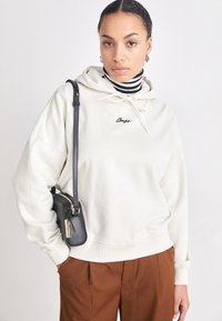 Hanorac crem cu șnur, logo brodat și manșete ribbate, asortat cu o geantă neagră tip crossbody și pantaloni maro, cu o țesătură texturată.