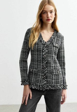Black and white tweed jacket with a v-neck, front buttons, and two pockets; features a textured pattern and ruffled cuffs.