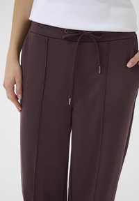 Pantaloni leggeri color bordeaux con chiusura a cordoncino, caratterizzati da pieghe laterali e una texture liscia. Ideali per un abbigliamento casual o rilassato.