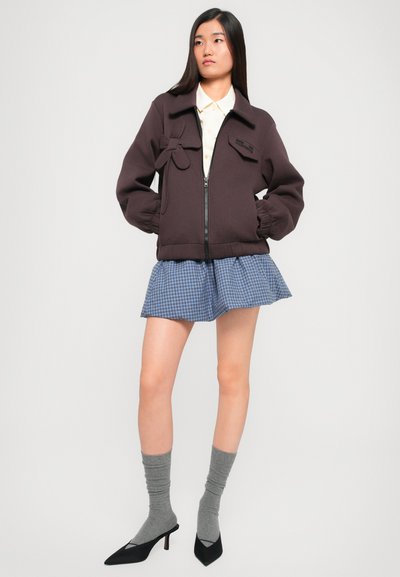 Καφε zip-up μπουφάν με χαλαρή εφαρμογή, ριμπ μανσέτες και λάδι. Συνδυασμένο με πλισέ φούστα, γκρι κάλτσες και μαύρα μυτερά τακούνια.