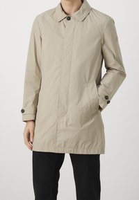 Kehittävä beige kevyt takki, jossa on kaulus, yksittäinen nappilukko, kaksi sivutaskua ja nappihaisari; valmistettu sileästä, teksturoidusta kankaasta.