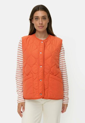 Woman wearing an orange quilted vest with snap buttons over a long-sleeve white shirt with red stripes and white pants.